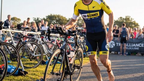 Triathlete in the yellow and navy Tri Alliance trisuit wheeling his bike out of transition past a cheering crowd