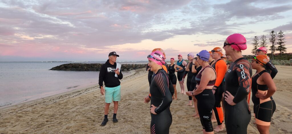 Tri Alliance coach talking a group of triathletes through an early morning beach swim session at sunrise