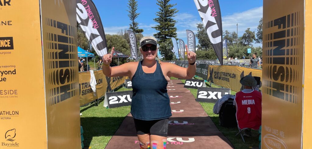 Smiling triathlete celebrating under the finish arch at a 2XU Triathlon Series race in Elwood
