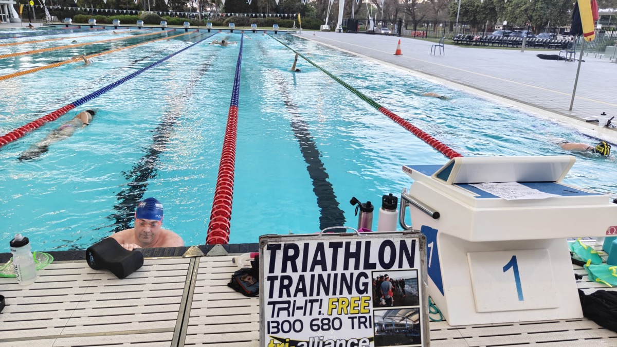 Smiling swimmer in a blue cap resting at the wall of the MSAC outdoor pool beside the Tri Alliance Triathlon Training sign