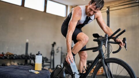 Triathlete on an indoor wind trainer reaching for a drink bottle, with a second bottle of sports drink and an energy gel on a towel beside the bike