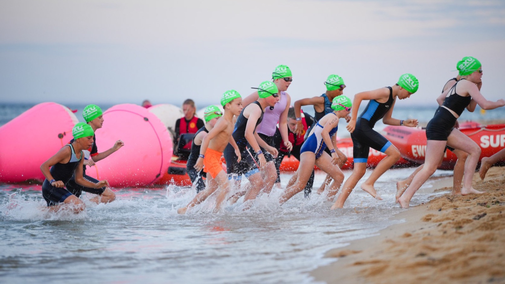 Triathletes entering the sea for a swim start with pink buoys and red boats in the background.