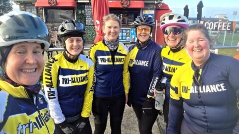 Six Tri Alliance women in club cycling kit smiling at a café stop on a group ride